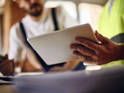 Close-up: Worker's hands in a safety vest hold/review a digital tablet; second worker is blurred behind.