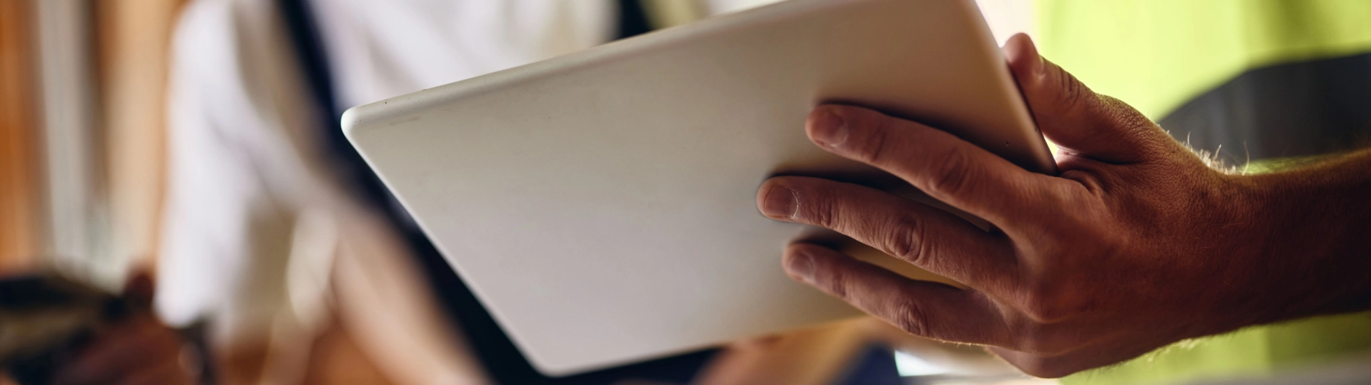 Close-up: Worker's hands in a safety vest hold/review a digital tablet; second worker is blurred behind.